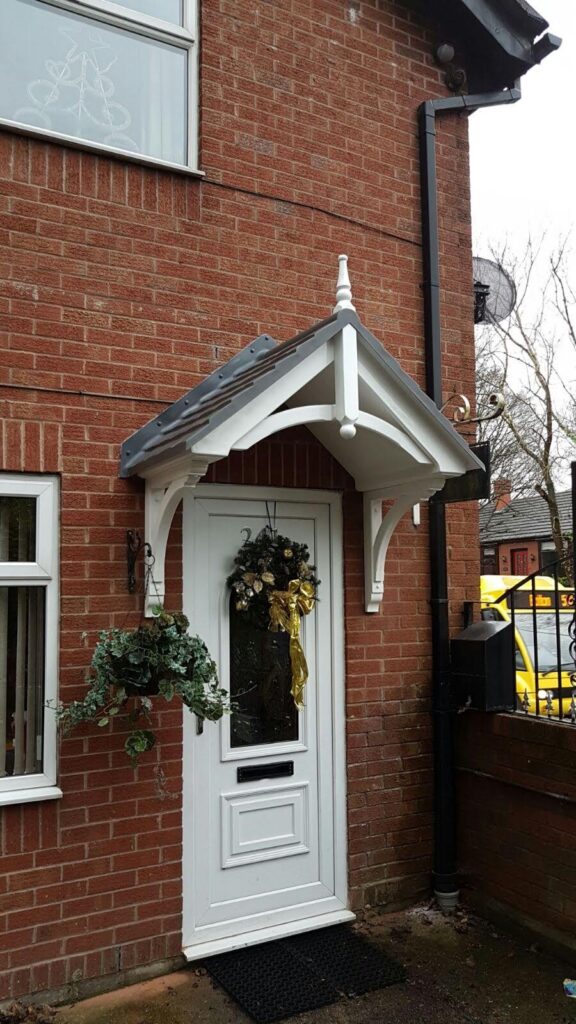 A duo pitch door canopy with a finial and curved brace feature, above a decorated white front door