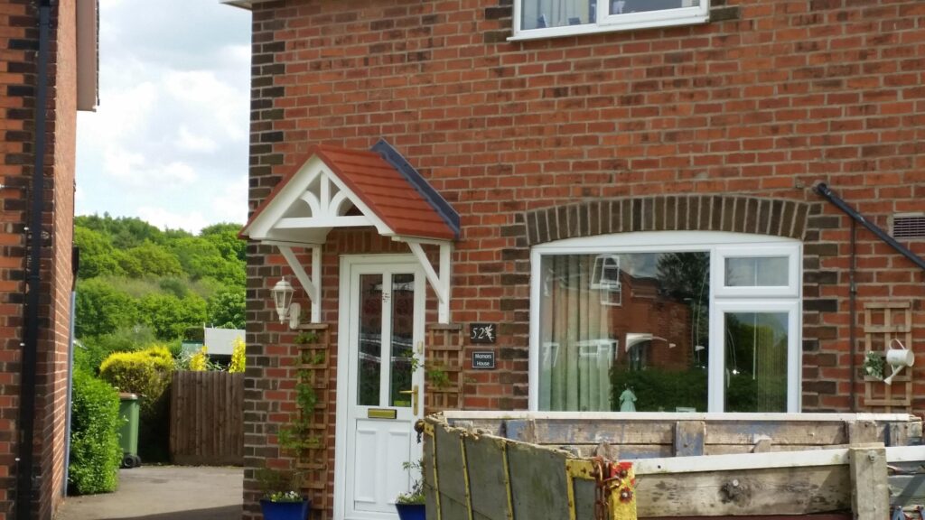 A white door canopy with a red roof above the front door of a house, with a skip in front